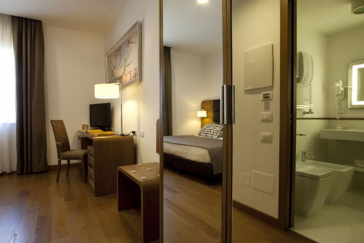A hotel room with a bed, a desk, a chair, a small table, a TV, a lamp, and a bathroom with a sink and mirror.