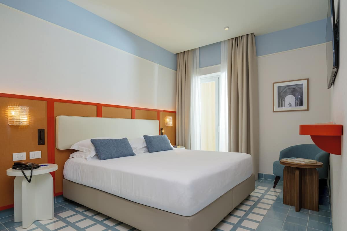 Una habitación de hotel con una cama grande, una mesita de noche con teléfono, una pequeña mesa redonda con un libro, una lámpara de pared y un cuadro en la pared.