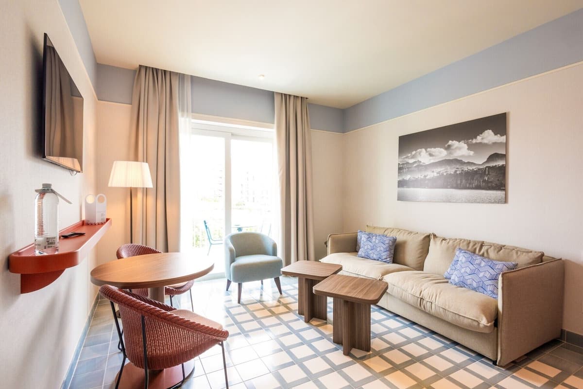 Habitación de hotel con sofá, dos sillones, una mesita, lámpara, televisor y ventana con cortinas.