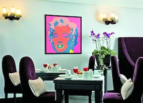 Un coin repas avec une table dressée pour deux personnes, un portrait de style pop art encadré et une plante en pot ornée de fleurs violettes.