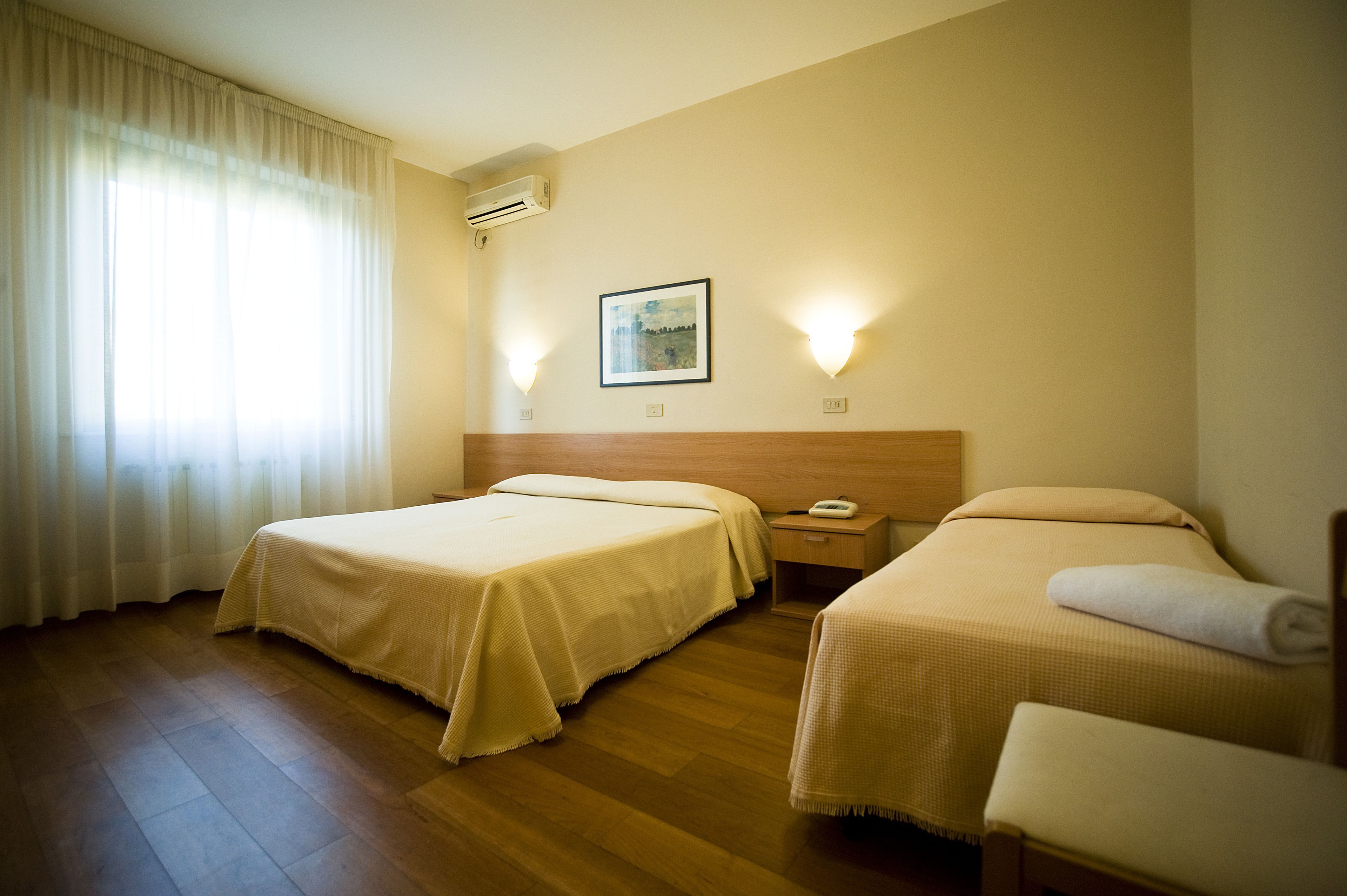 A hotel room with two beds, a wooden floor, a nightstand, and a wall-mounted air conditioner.