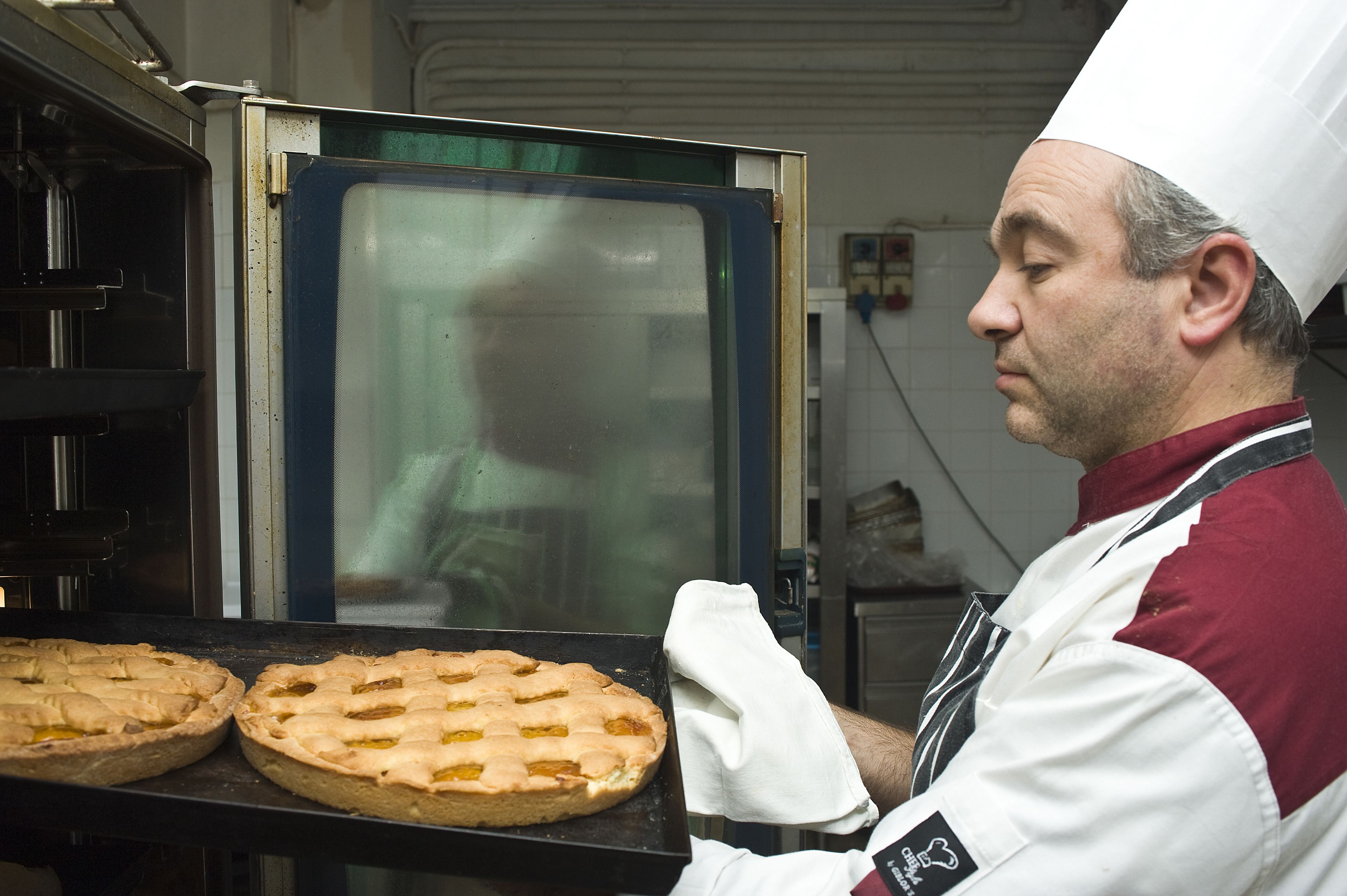 Un chef con uniforme blanco y rojo y un alto gorro blanco, preparando tartas en una cocina industrial con electrodomésticos de acero inoxidable y un horno grande.