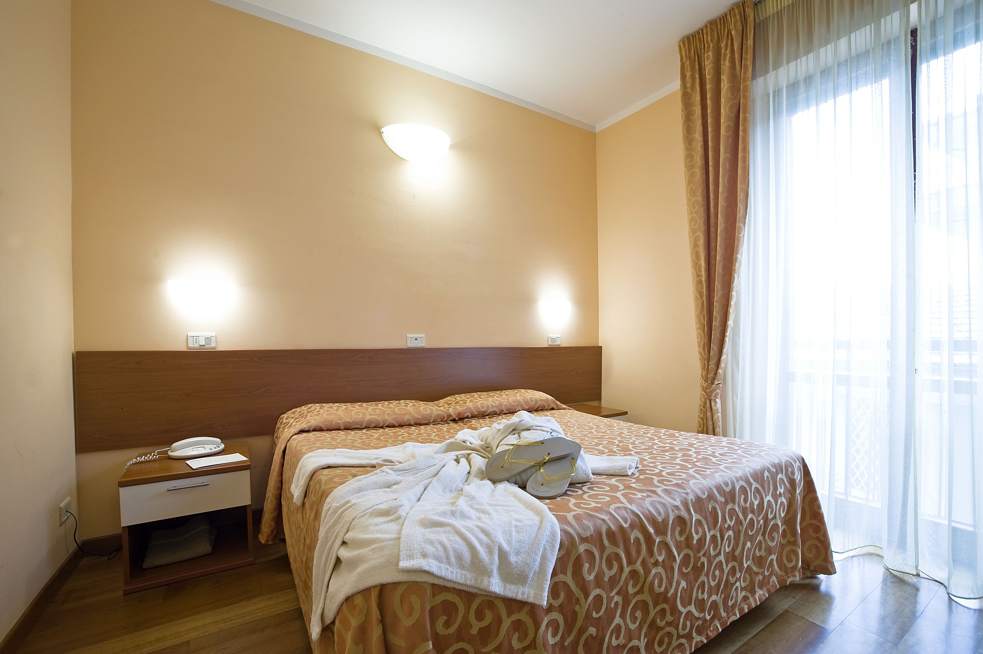 A hotel room with a bed, a nightstand with a phone, and a window with curtains.