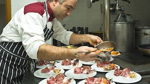 Un cuiner preparant aliments en una cuina amb electrodomèstics d'acer inoxidable i parets de rajola blanca.