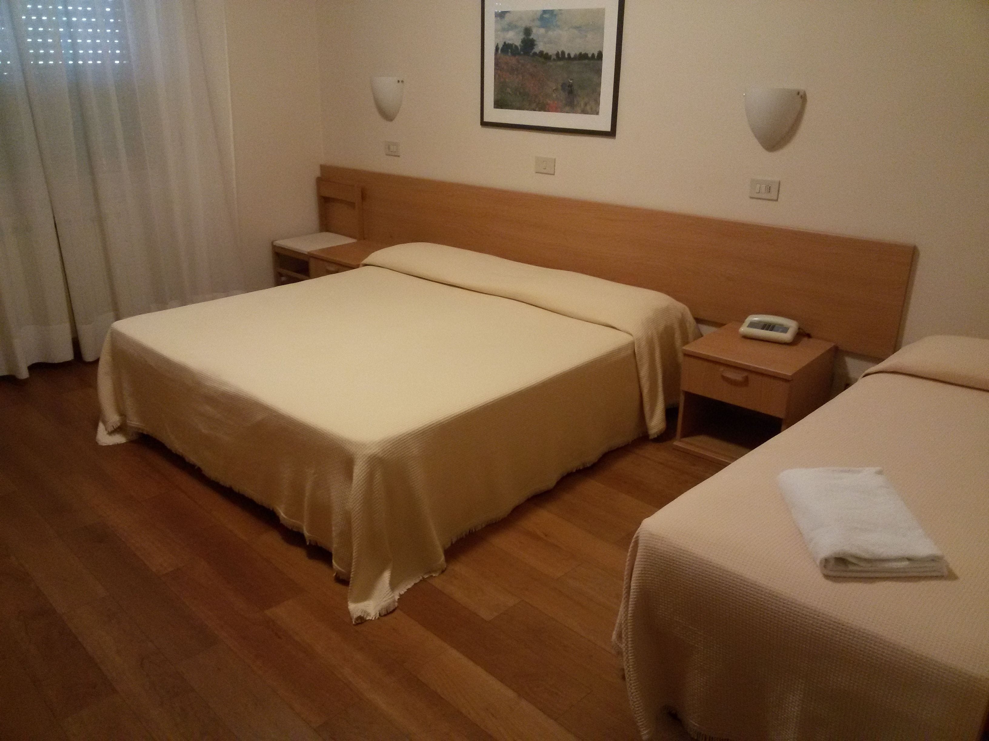 A hotel room with a double bed, a single bed, a bedside table with a lamp, and a wall-mounted telephone.