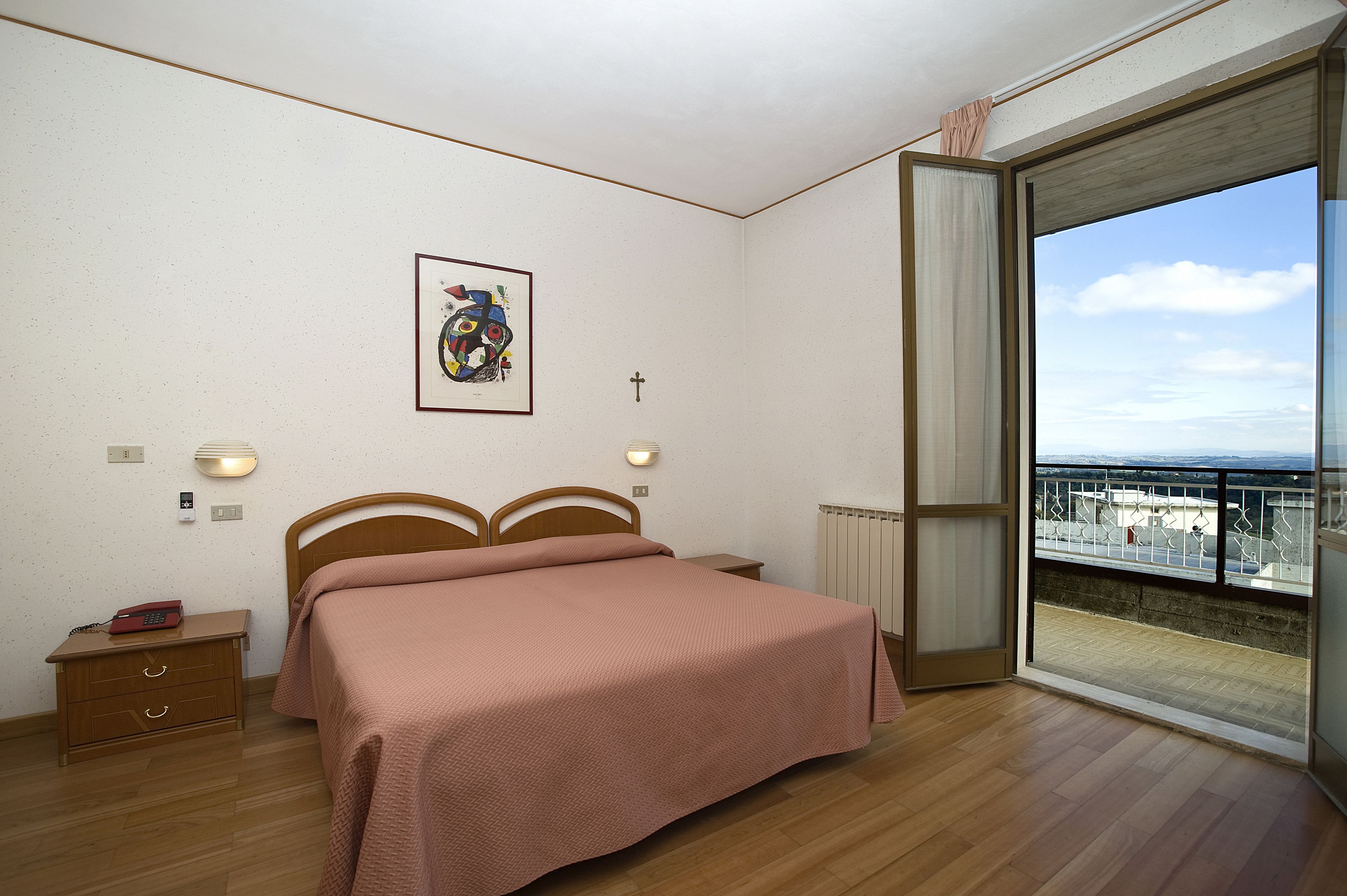A bedroom with a bed, bedside tables, a framed picture, a cross, and a balcony with a view.