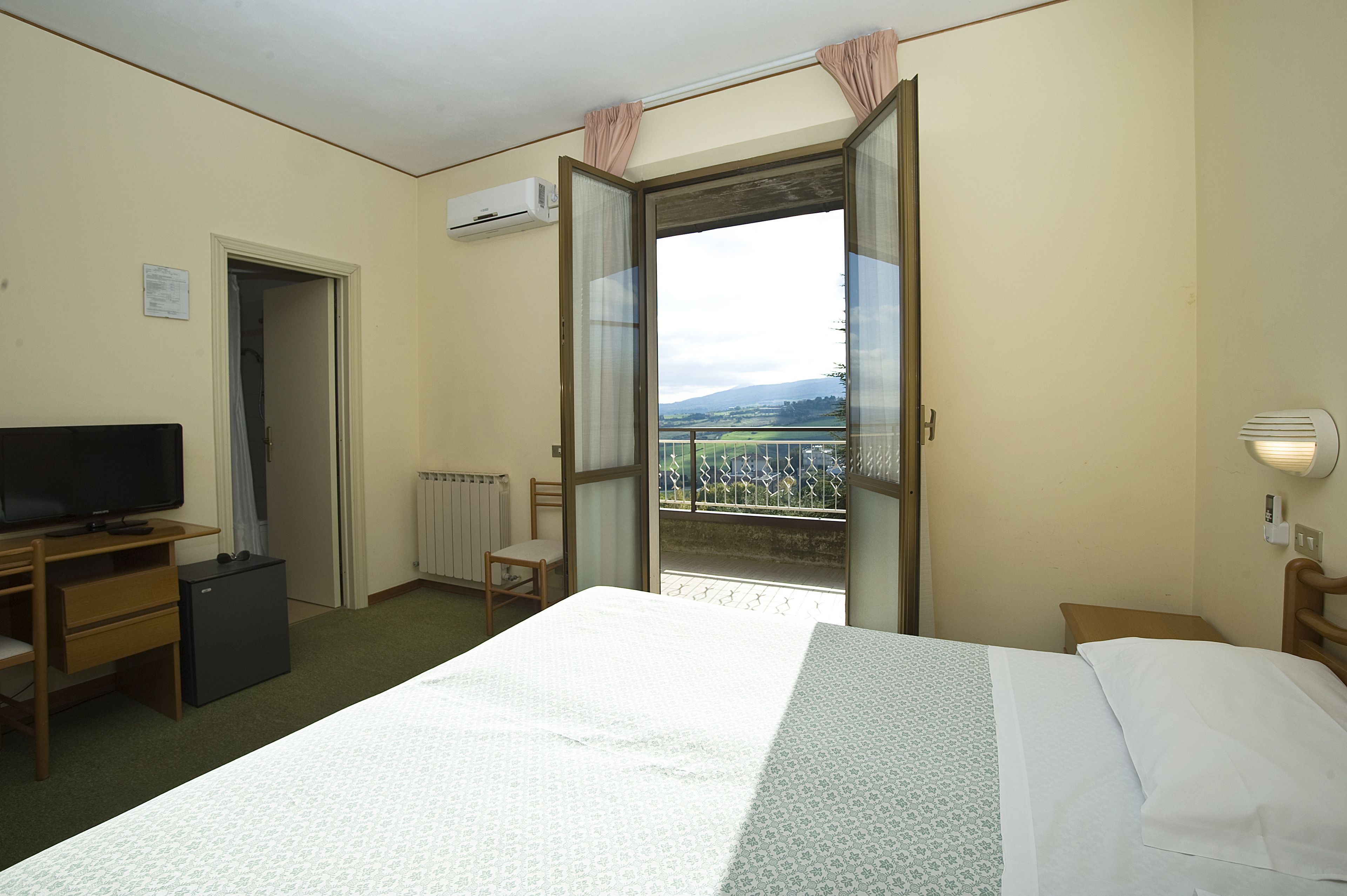 Ett hotellrum med en säng, ett skrivbord med en tv, ett litet kylskåp, en stol och ett fönster med utsikt över en balkong och berg.