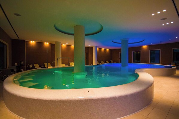 A large indoor swimming pool with a circular design, surrounded by lounge chairs and a tiled floor.