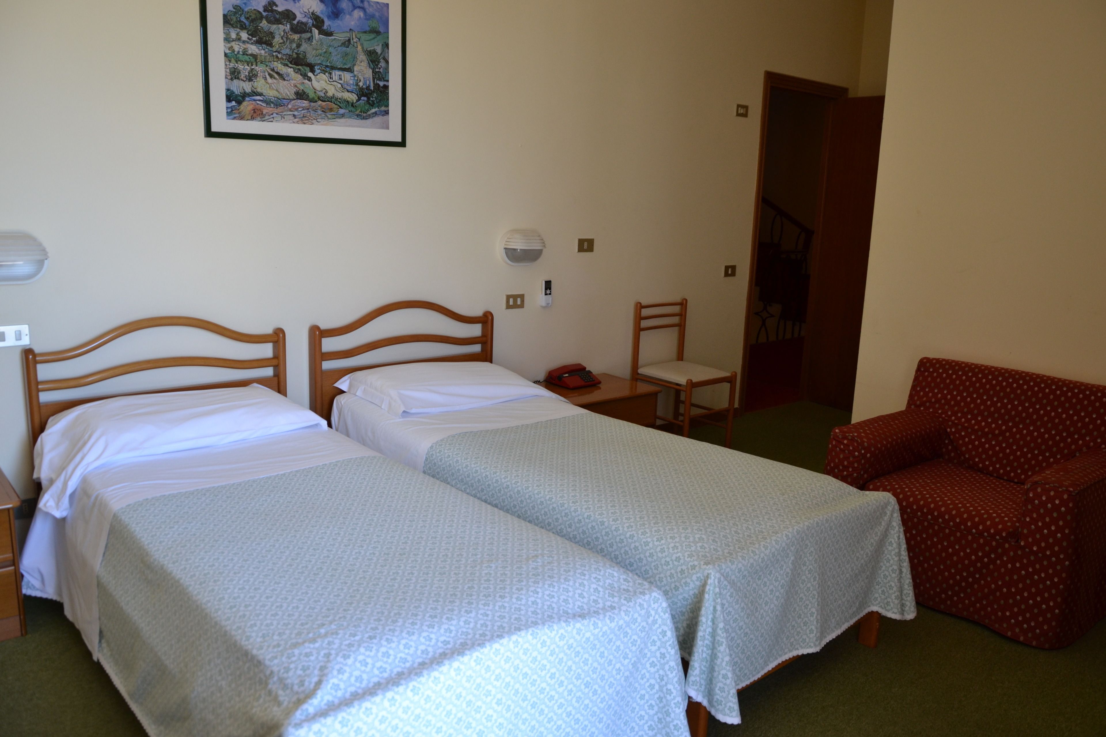 A hotel room with two single beds, a red armchair, a painting on the wall, and a staircase in the background.