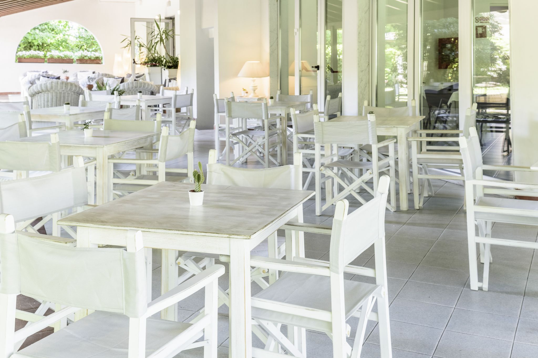 A spacious outdoor dining area with white wooden tables and chairs, surrounded by glass walls and potted plants.