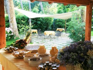 Un área de comedor al aire libre con mesas y sillas, rodeada de vegetación y un espacio de estar techado.