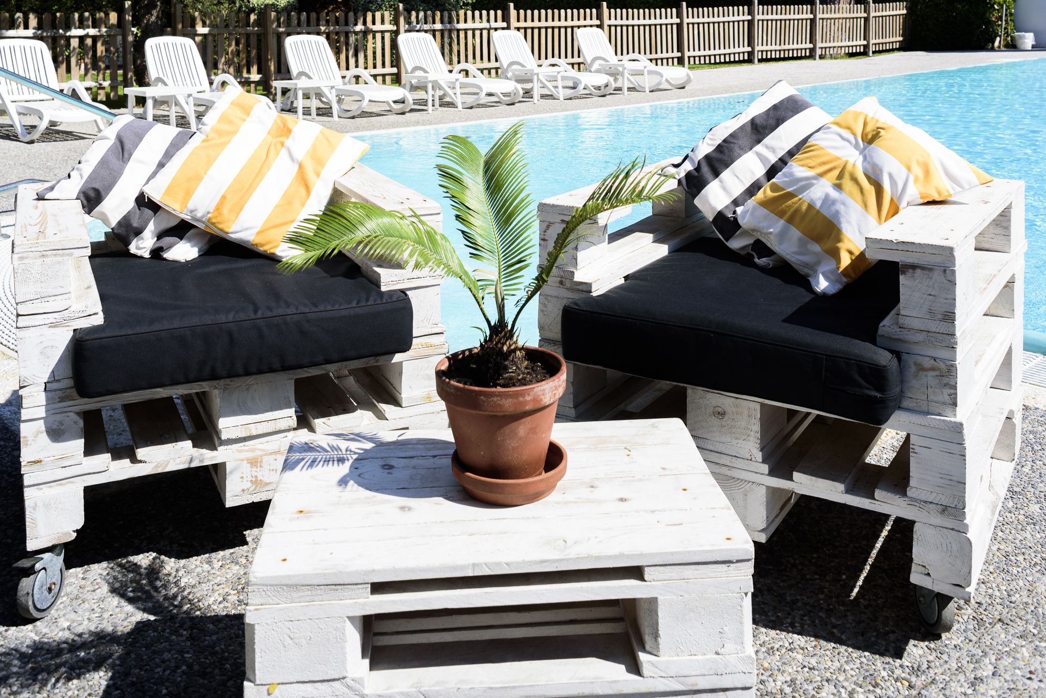 Dos sillones de descanso con cojines negros y almohadas a rayas amarillas y blancas, una planta en maceta y una mesita baja frente a una piscina.