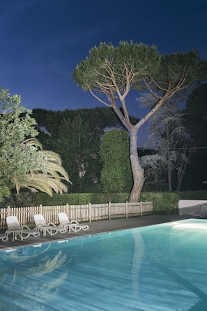 Una piscina con sillas de descanso y un árbol grande al fondo.