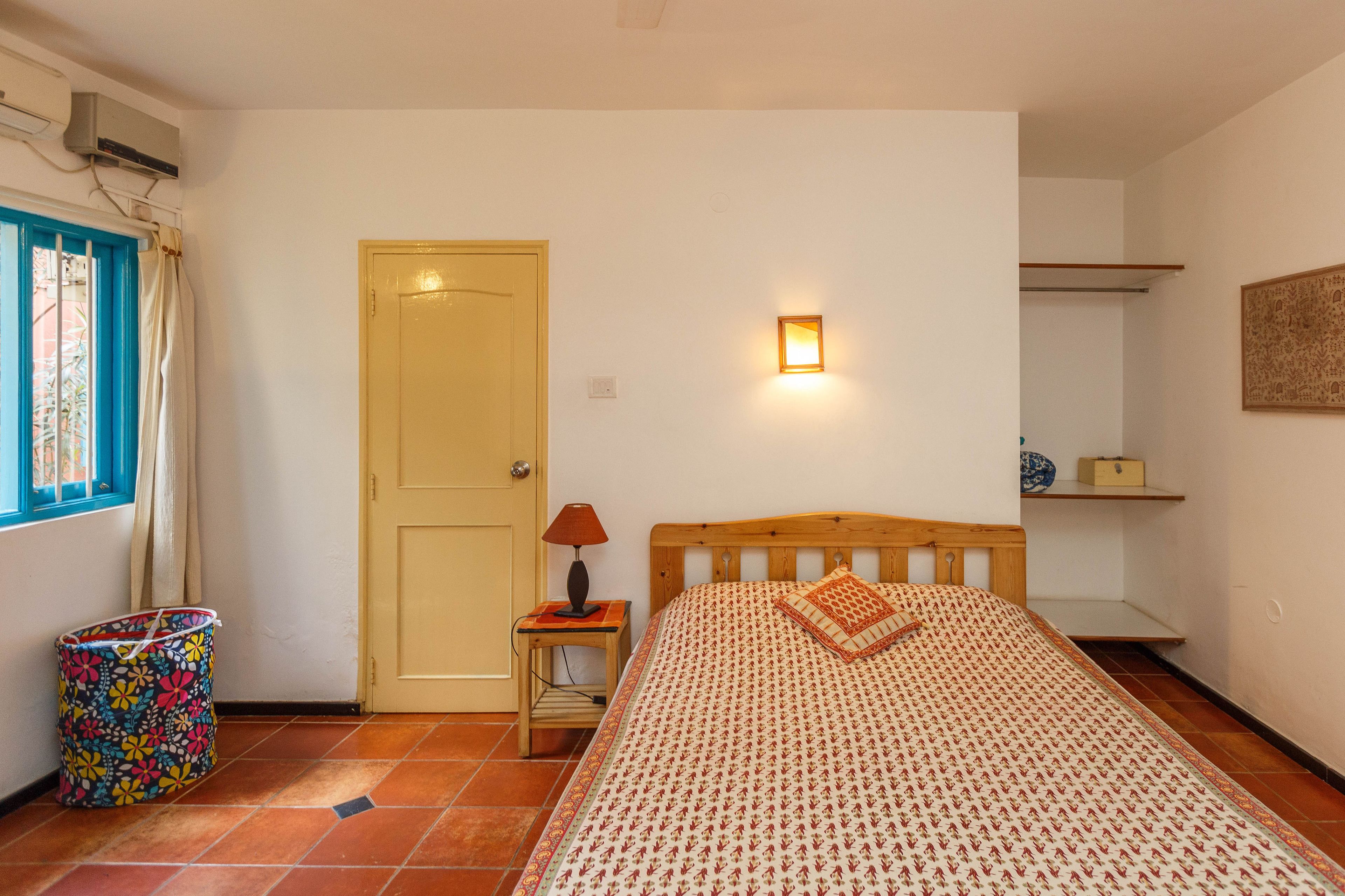 Un dormitorio con cama, una mesita de noche con lámpara, una ventana con cortinas, una puerta y un pequeño estante con objetos decorativos.