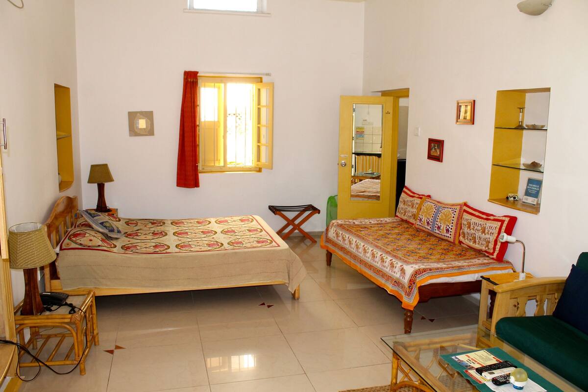 A room with two beds, a chair, a table, a lamp, a window with curtains, and a mirror.