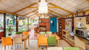 A restaurant with a covered patio, wooden tables and chairs, a kitchen area, and a view of the outside.