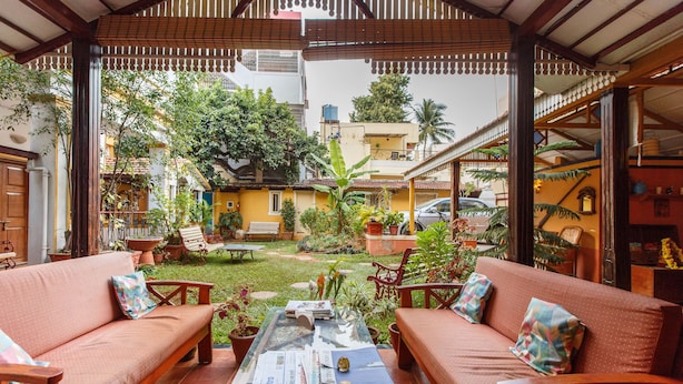 A covered outdoor seating area with wooden furniture and a glass table, surrounded by potted plants and a view of a residential neighborhood.