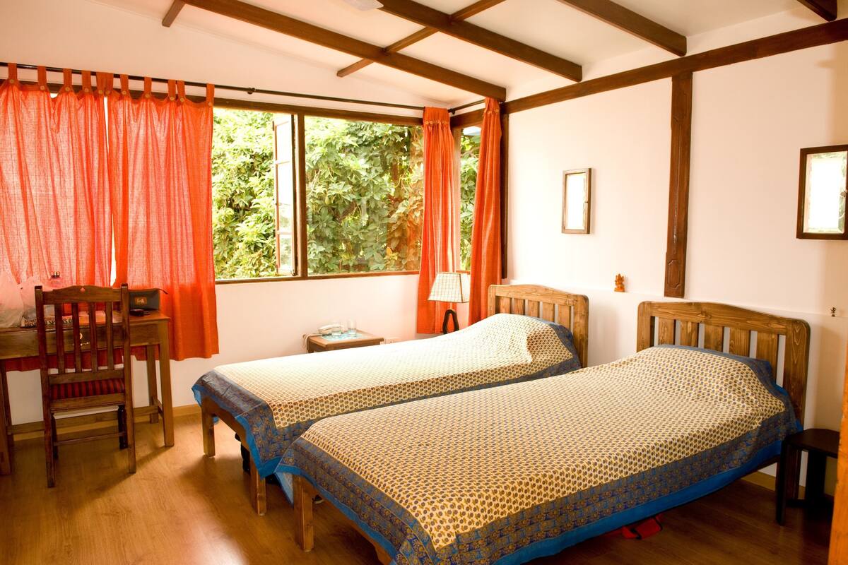 A room with two single beds, a wooden desk with a chair, a small table with a lamp, and a window with curtains.