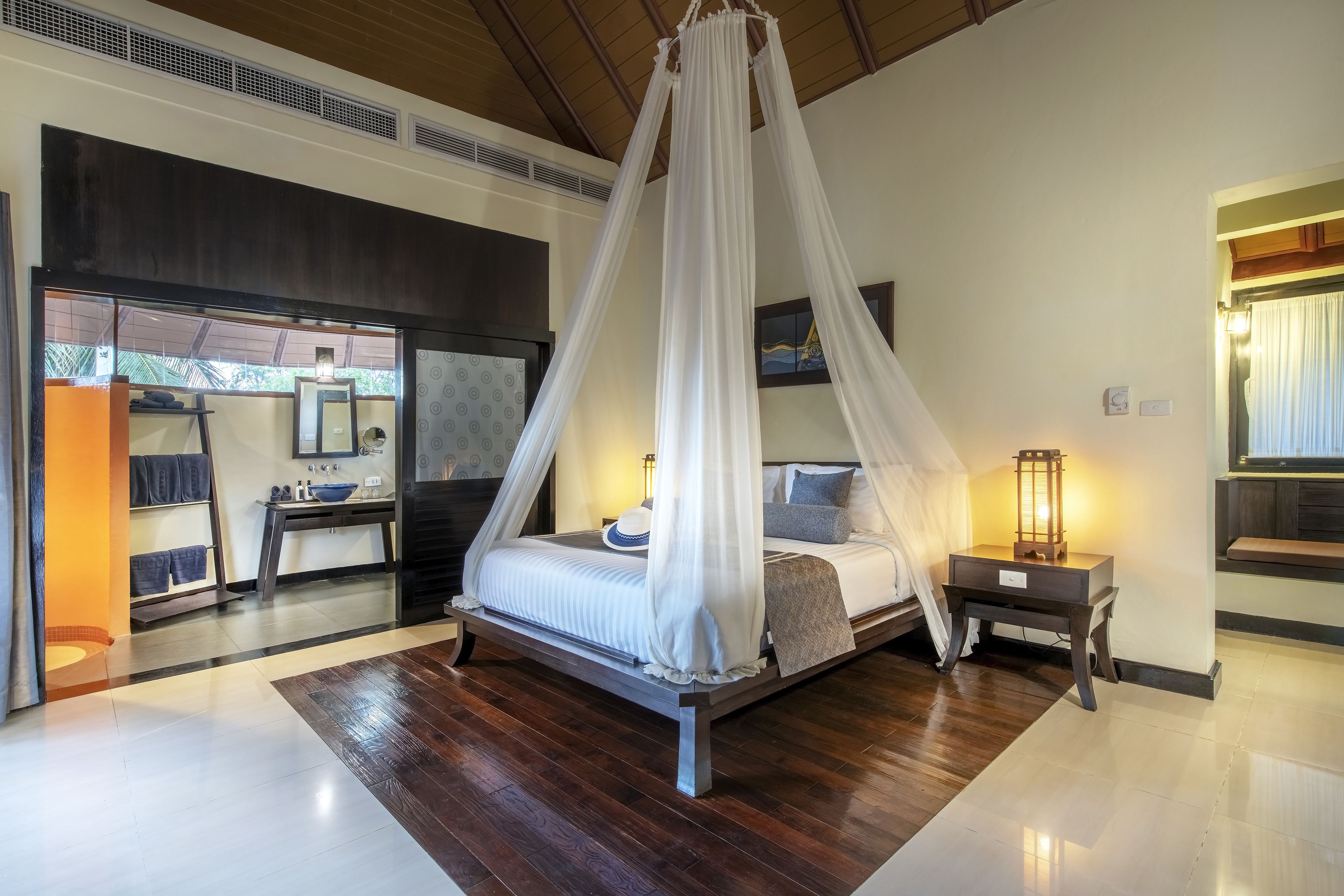 A bedroom with a canopy bed, a bedside table with a lamp, and a view of the outside.