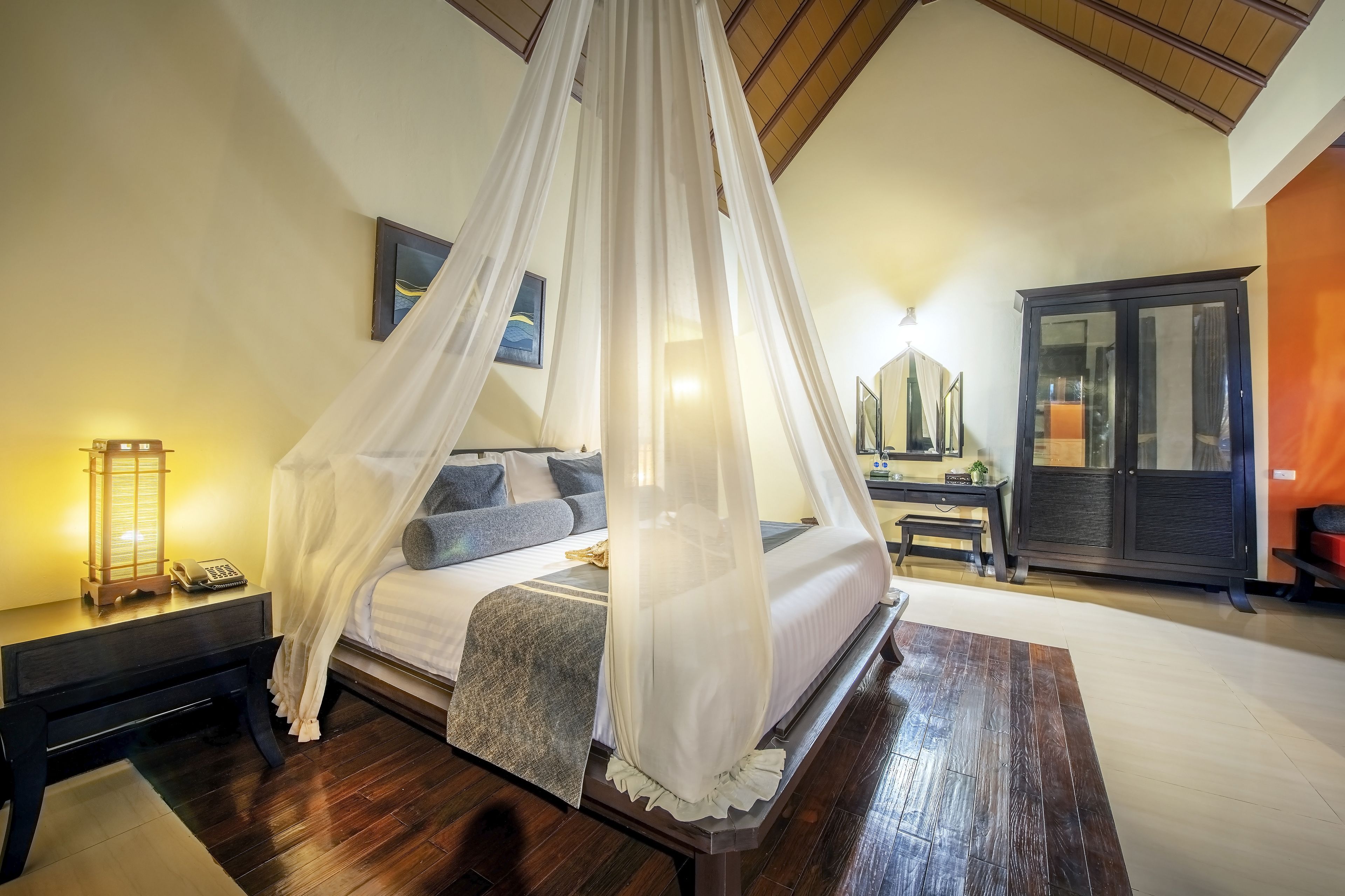 A bedroom with a canopy bed, a nightstand with a lamp, a wardrobe, and a desk with a chair.