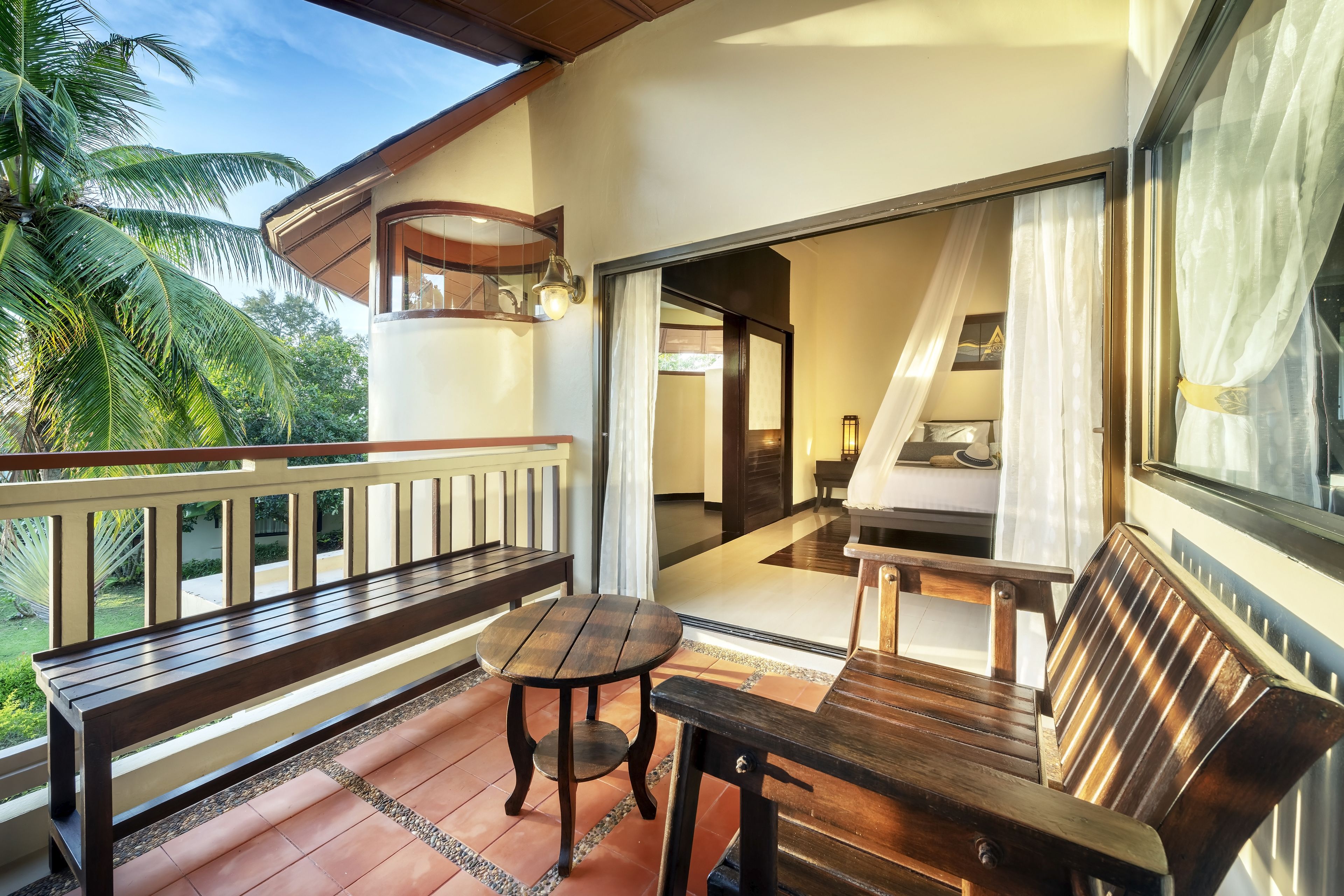 Un balcón con muebles de madera, incluyendo un banco, una mesa y sillas, con vista a un jardín con una palmera.