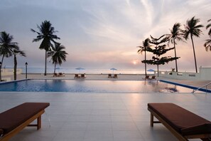 Área de piscina con sillones de descanso y sombrillas, palmeras y vista al océano al atardecer.