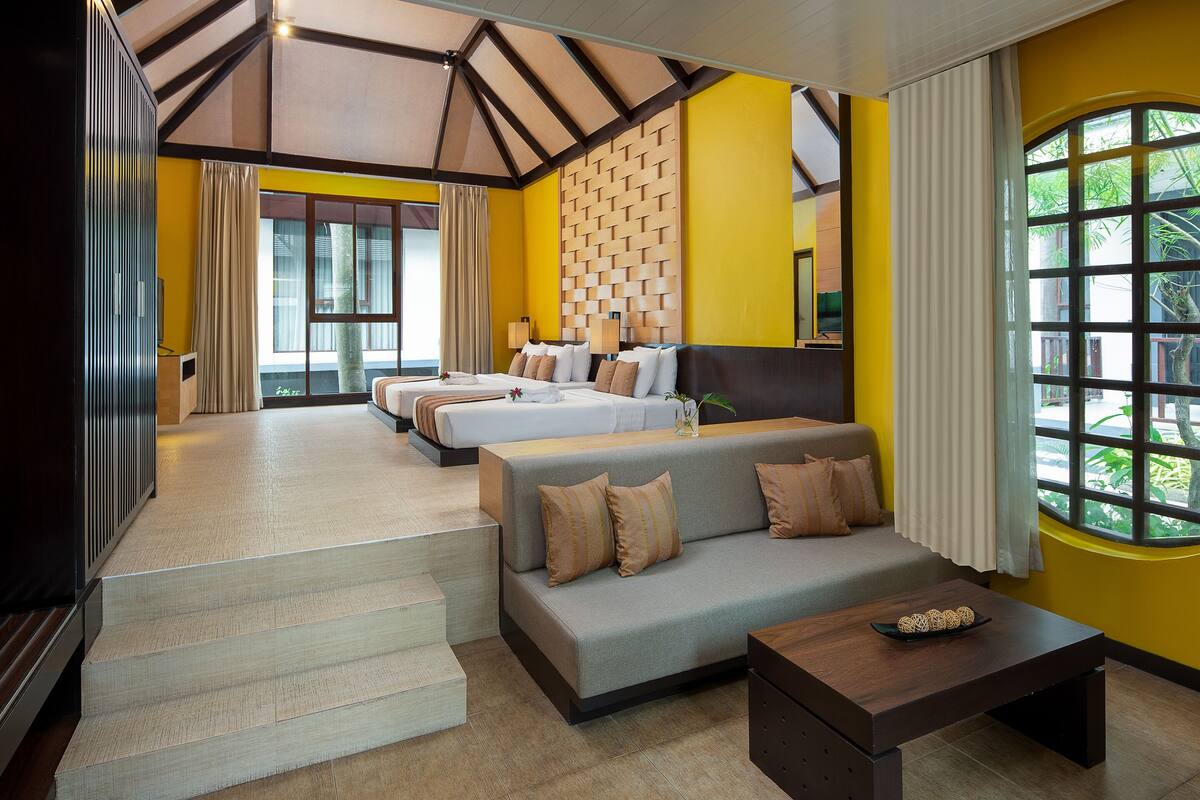 A hotel room with a sofa, a bed, a coffee table, and a view of greenery through a window.