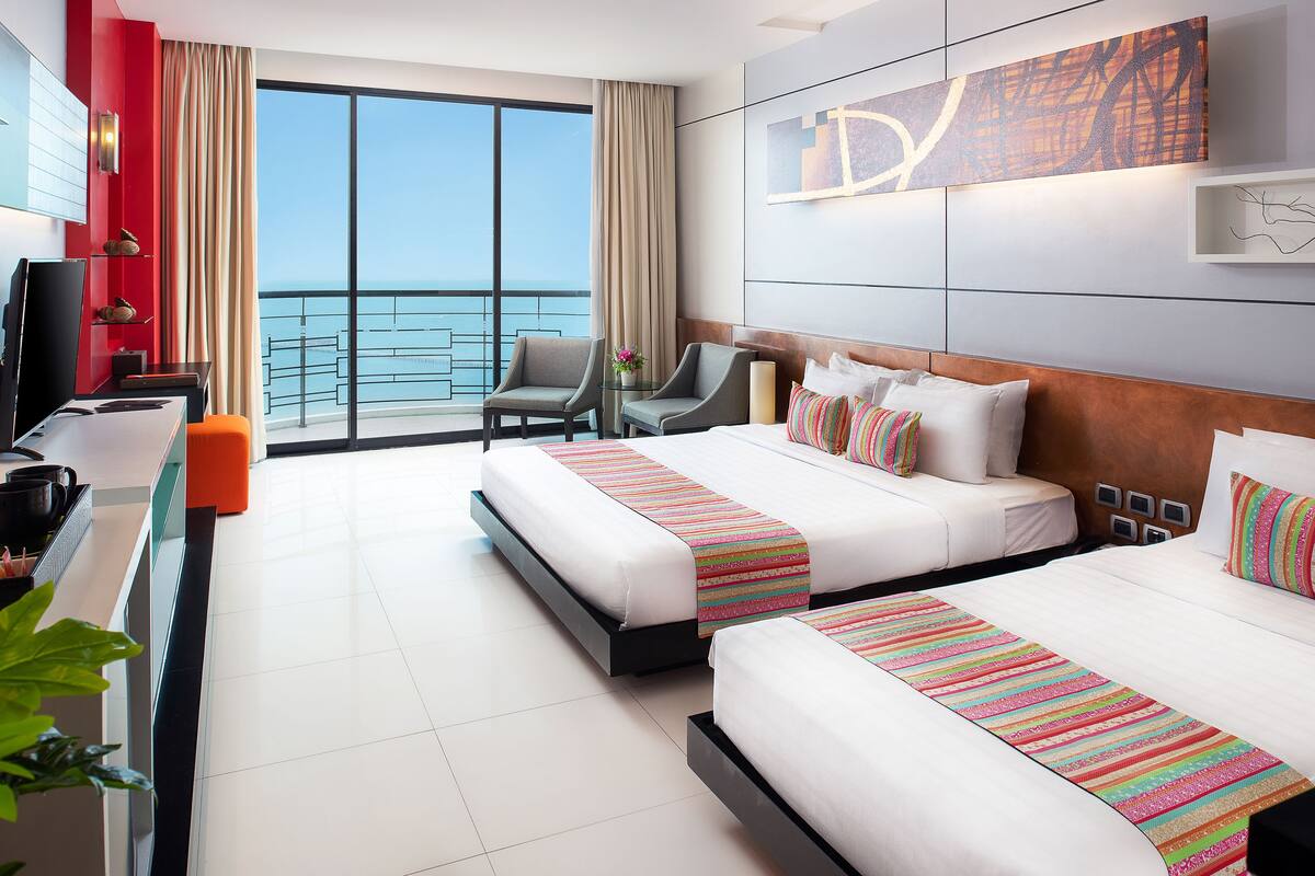 A hotel room with two beds, a desk, a chair, and a large window with a view of the sea.