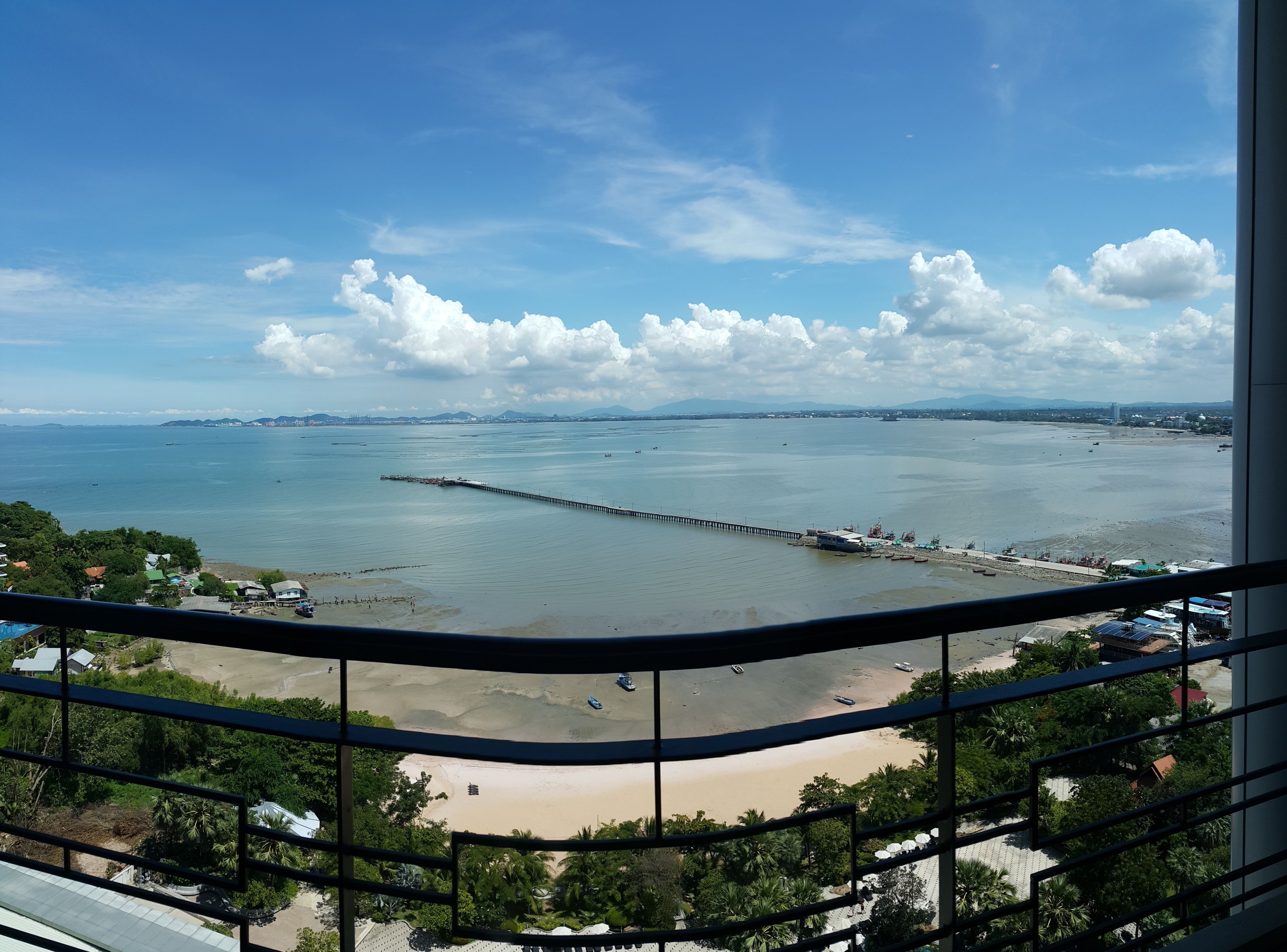 A coastal view from a balcony with a pier extending into the water, a beach, and distant mountains.
