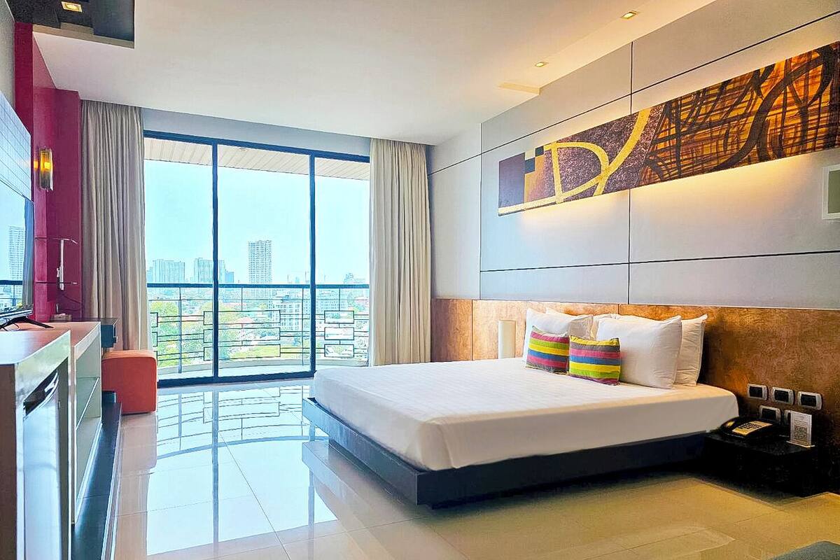 A modern hotel room with a large bed, a desk, and a view of the city through floor-to-ceiling windows.