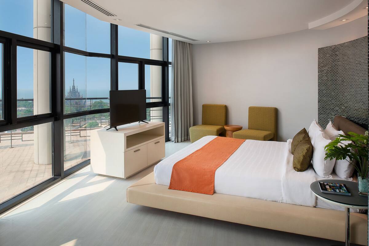 A modern hotel room with a large bed, a desk, two chairs, and a view of a temple through large windows.