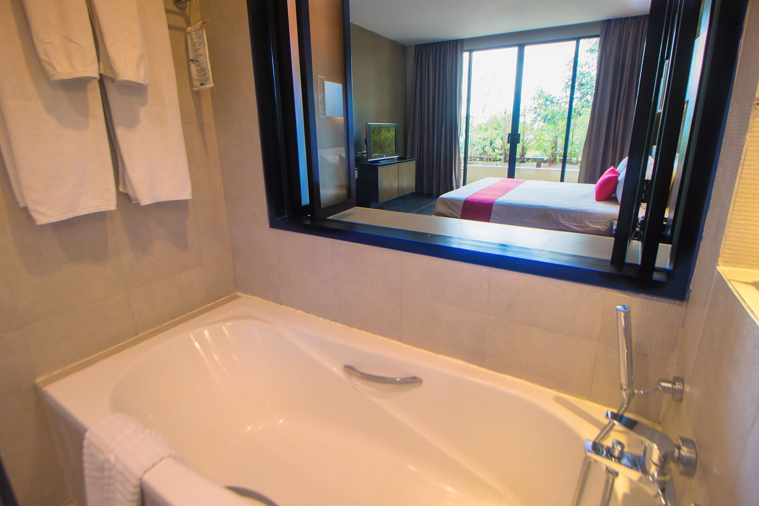 A hotel bathroom with a large bathtub, a mirror reflecting a bedroom with a bed and curtains, and a towel rack with towels.