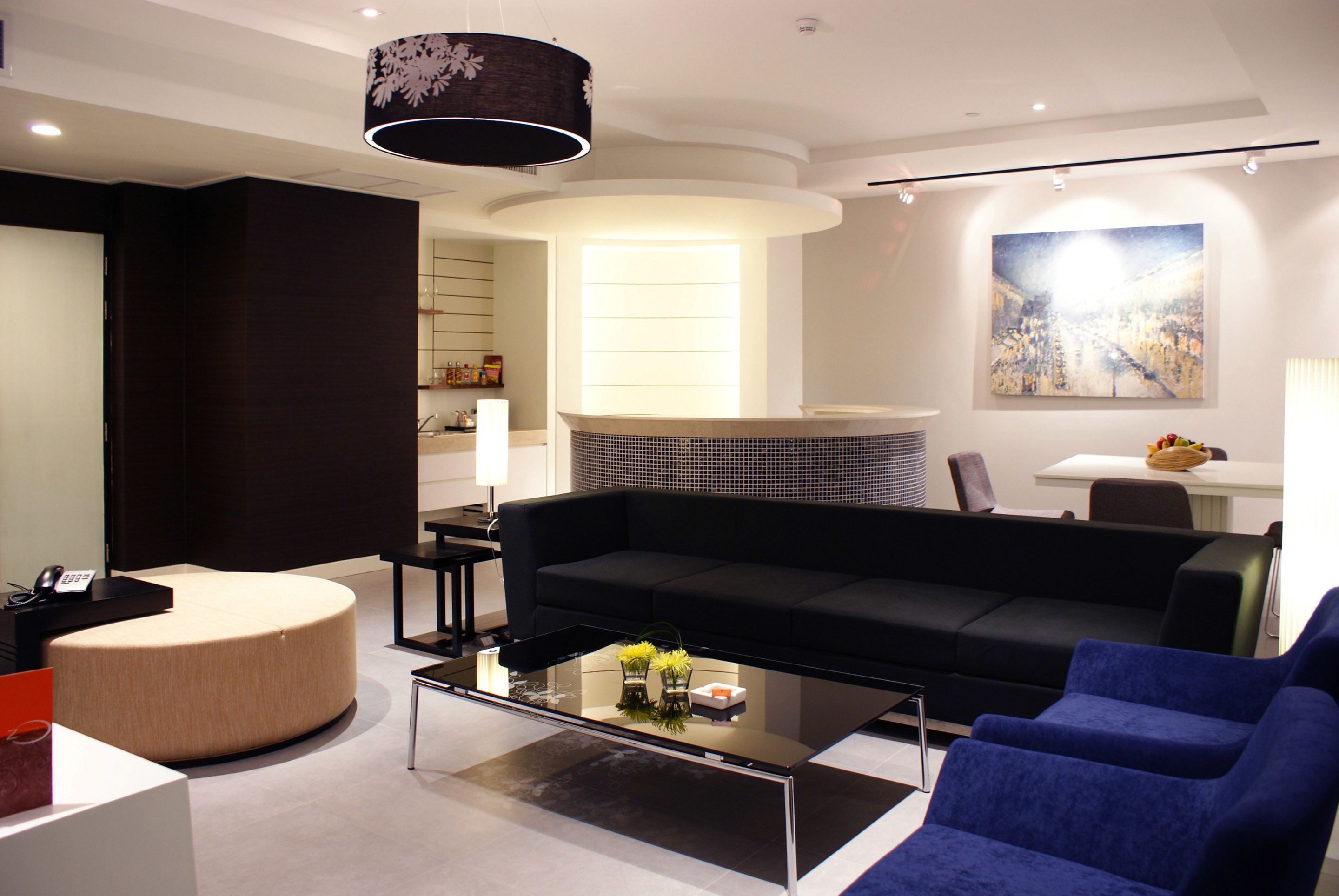 A modern living room with a black sofa, a round ottoman, a glass coffee table, and a large abstract painting on the wall.