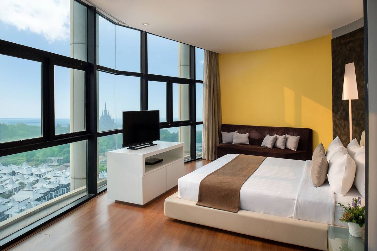 A modern bedroom with a large bed, a TV on a white console, a sofa, and a view of a cityscape through large windows.
