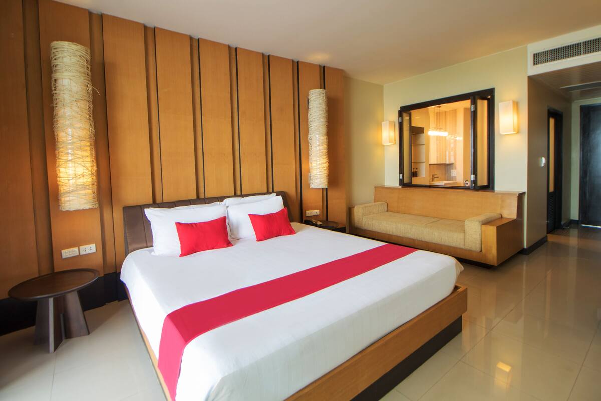 A large bed with white and red bedding, a wooden headboard, and a red accent strip. There is a beige sofa and a small round table beside the bed.