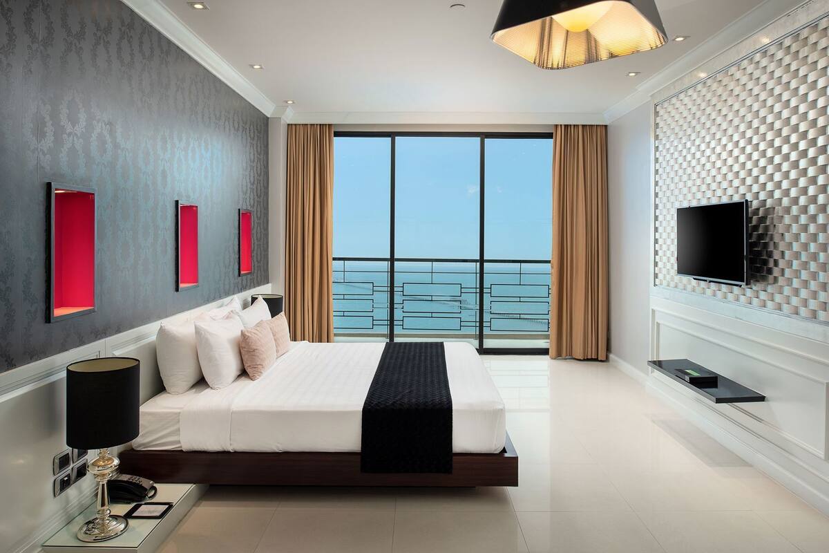 A modern bedroom with a large bed, a TV, and a balcony with a view of the sea.
