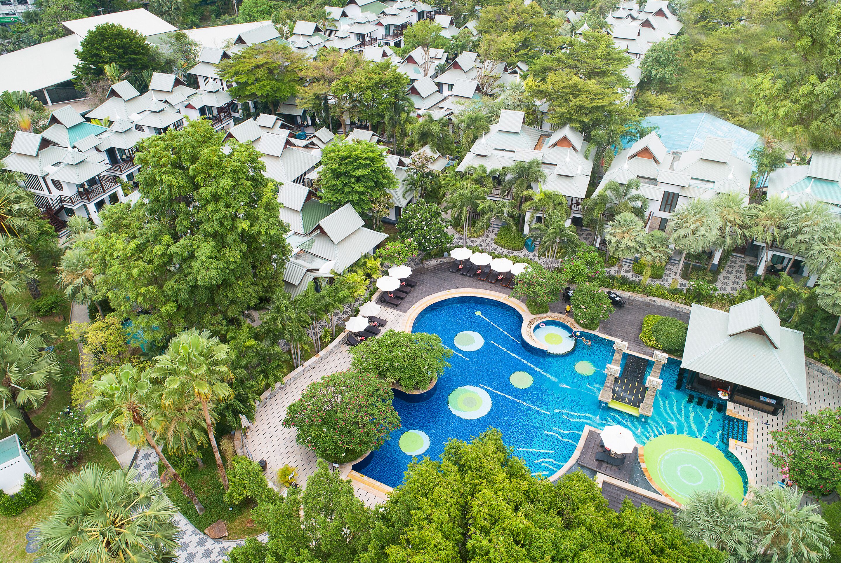 Eine Vogelperspektive zeigt ein Resort mit mehreren bungalowsartigen Unterkünften, einem zentral gelegenen Schwimmteich mit kreisförmigem Design und üppiger, umgebender Vegetation.