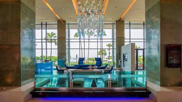 A modern lobby with a glass pool, seating area, and large windows offering a view of palm trees and a cityscape. - The Zign Hotel (Pattaya)