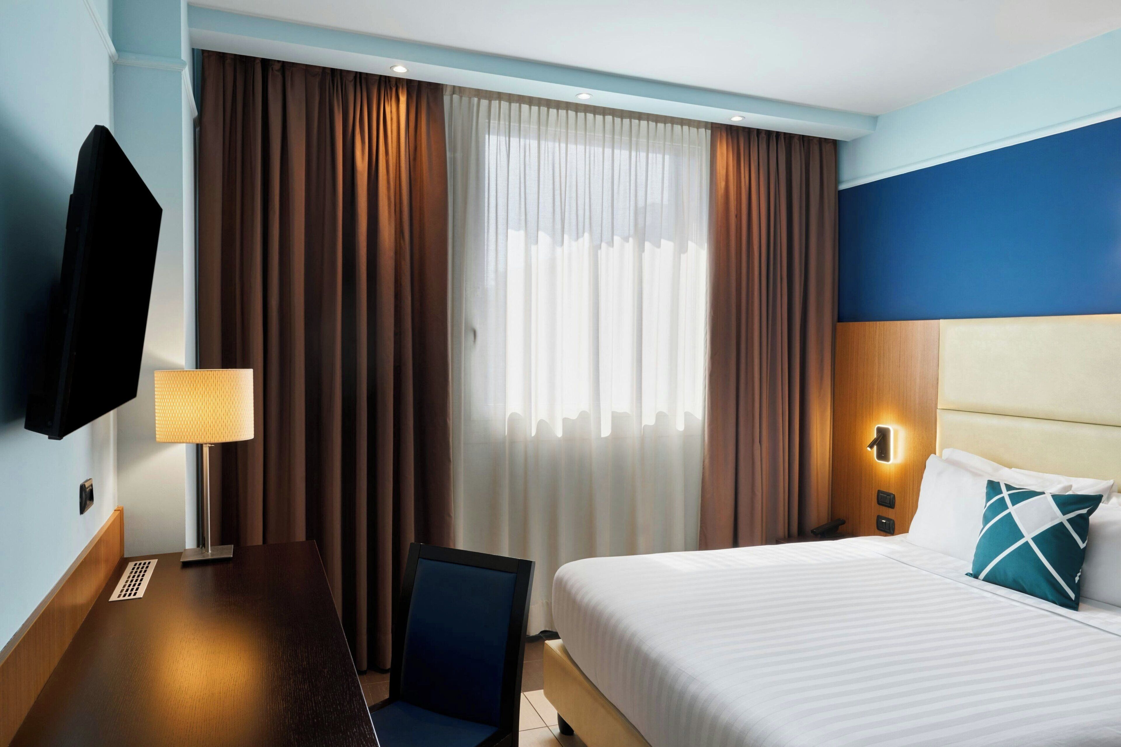 A hotel room with a bed, a desk, a chair, a TV, and curtains.