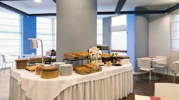 Un'area buffet con diversi cibi e una postazione caffè, circondata da tavoli e sedie in un ambiente moderno.