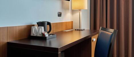 Habitación de hotel con escritorio de madera, una silla, televisor de pantalla plana montado en la pared, lámpara y ventana con cortinas.