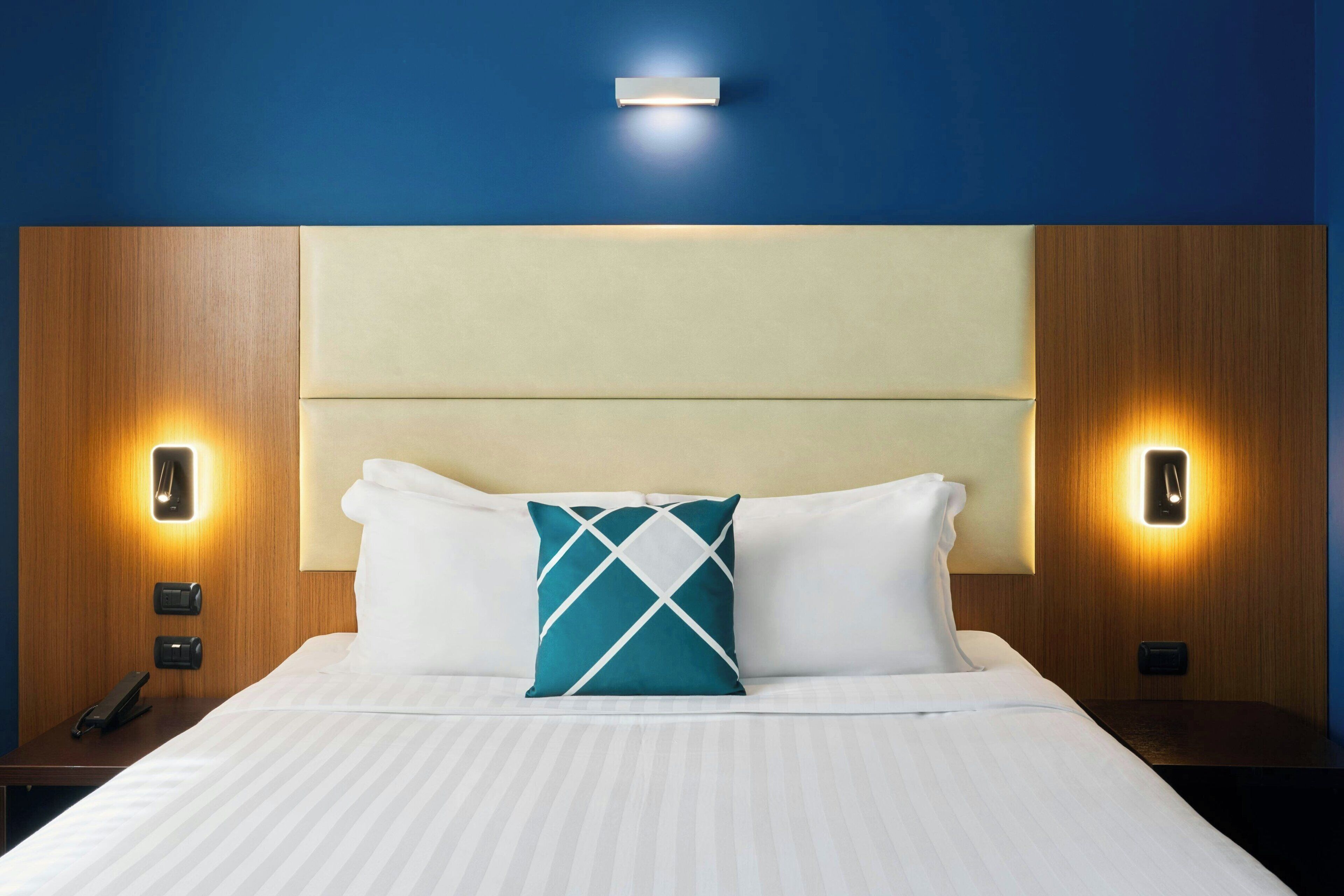 A hotel room with a large bed, two bedside tables, a headboard, and wall-mounted lighting.