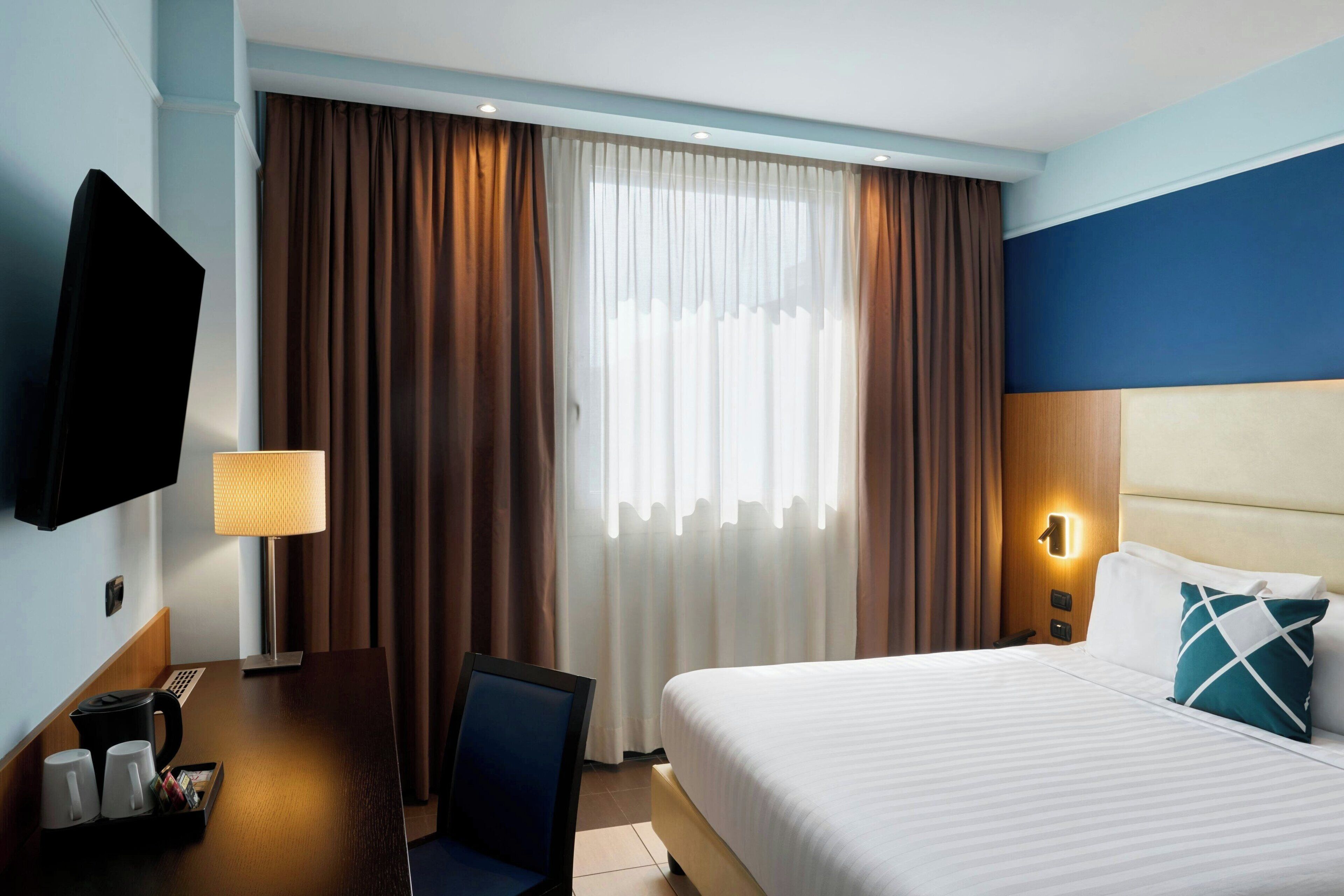 A hotel room with a bed, a desk with a lamp, a TV, and curtains.