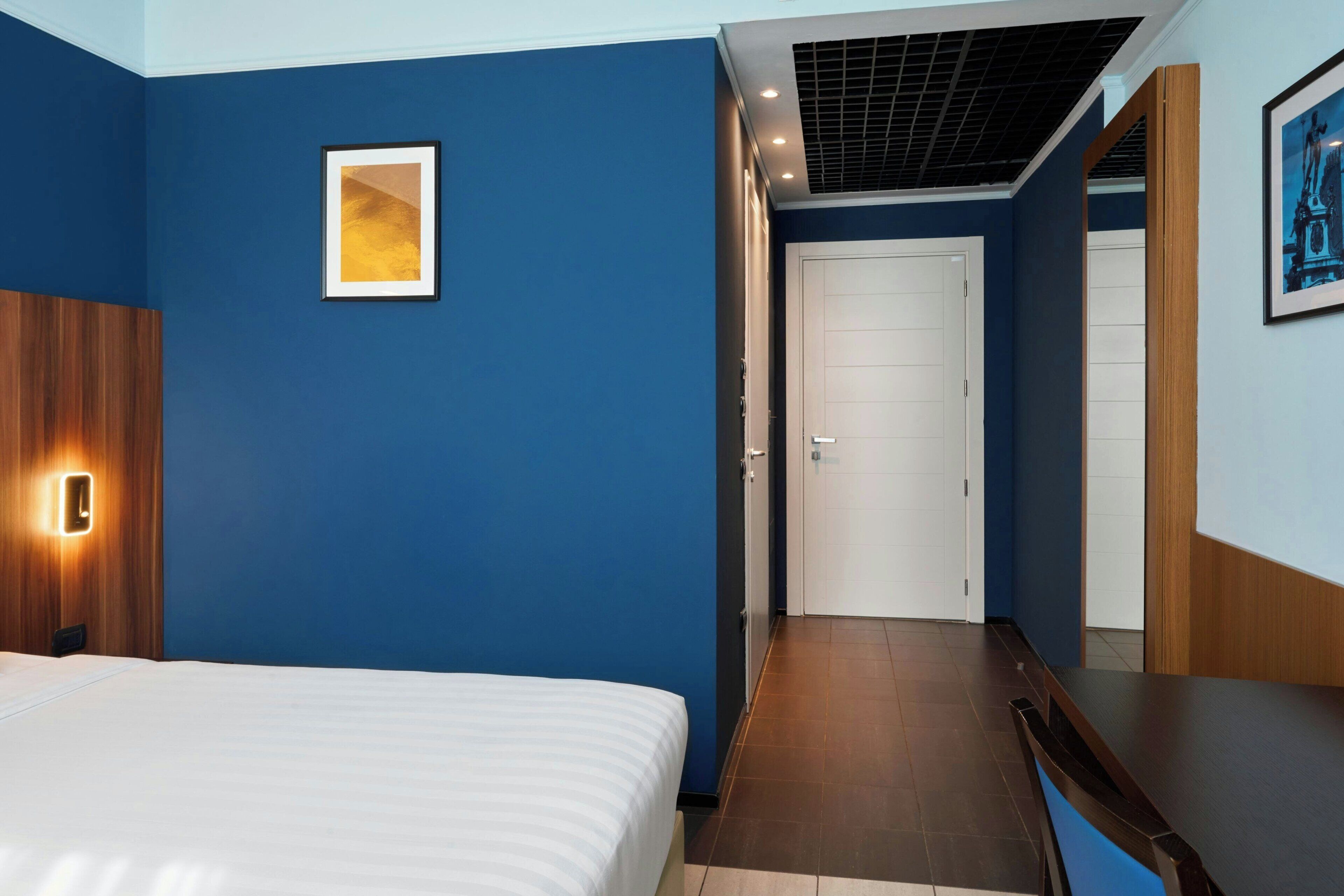 A hotel room with a bed, a door, and a hallway leading to another room.