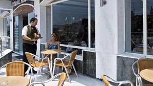 Un área de estar al aire libre con mesas y sillas redondas, un camarero sosteniendo una bandeja y un amplio ventanal con vistas a la ciudad.