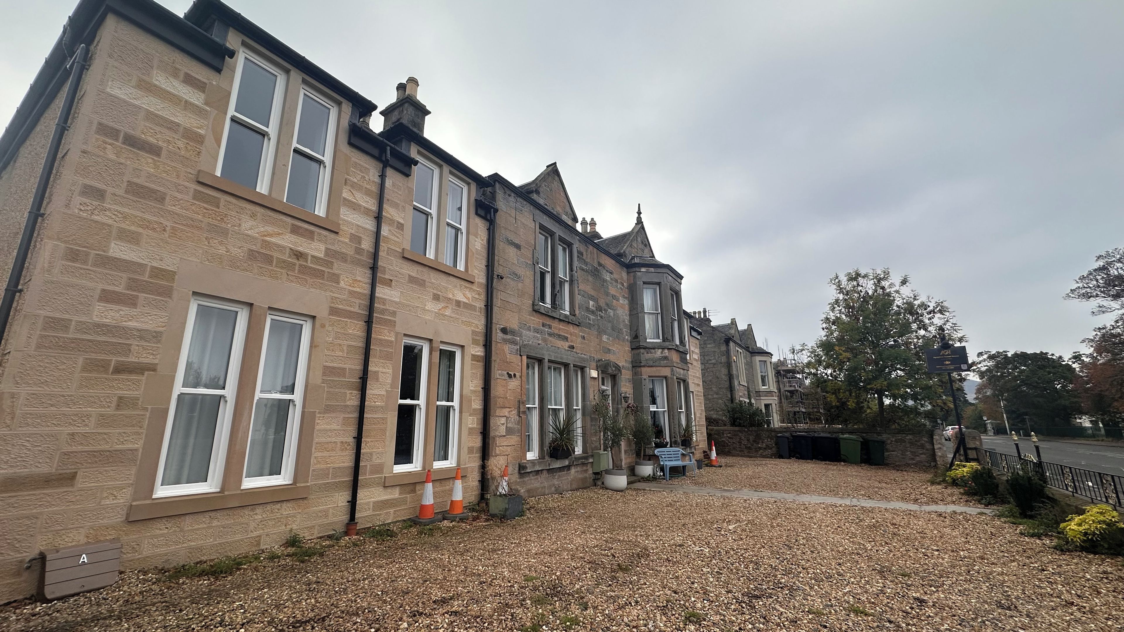 A row of stone buildings with multiple windows, a gravel driveway, and a small garden area with plants and a bench.