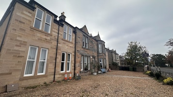 A row of stone buildings with multiple windows, a gravel driveway, and a small garden area with plants and a bench.