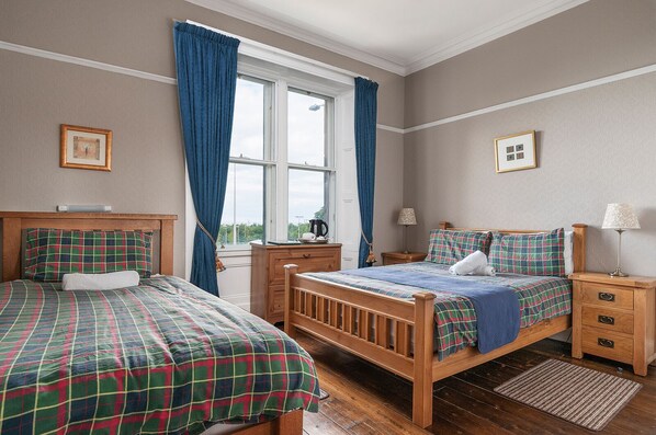Une chambre avec deux lits simples, une commode en bois, une table de chevet, une fenêtre avec des rideaux bleus et une vue sur l’extérieur.