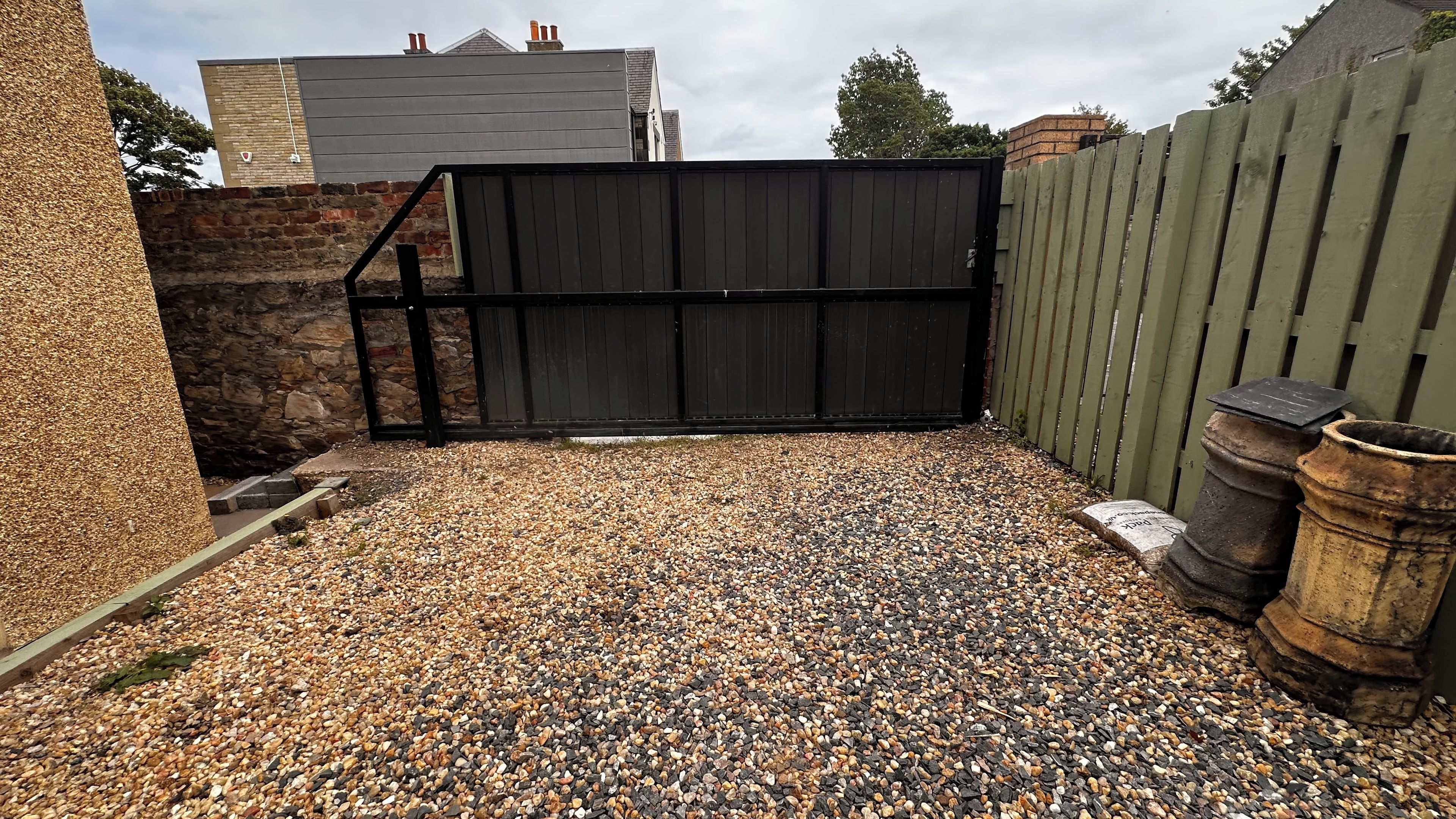 A gravel driveway leading to a black gate, surrounded by a stone wall and a wooden fence.
