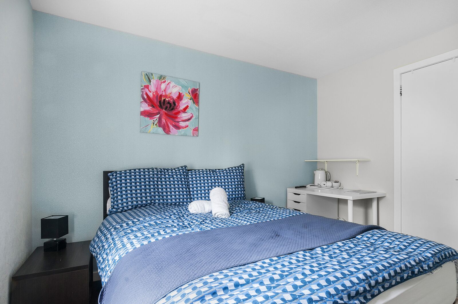 A neatly made bed with a blue and white patterned bedspread, a pillow, and a rolled towel on the bed. There is a small bedside table with a lamp and a book. A painting of a flower is on the wall above the bed.